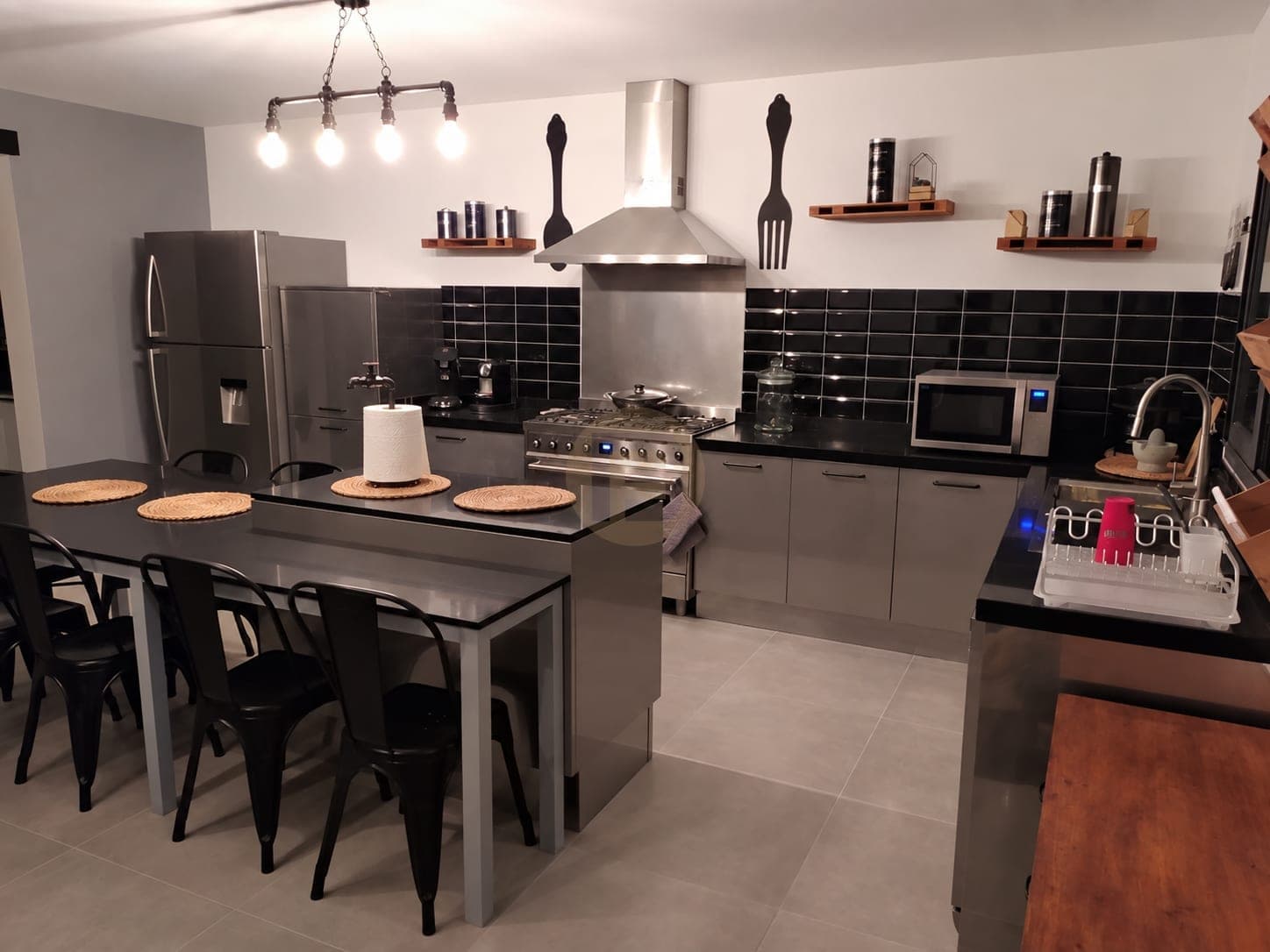 Stainless kitchen with a tall fridge column, range cooker and island table from the dining side